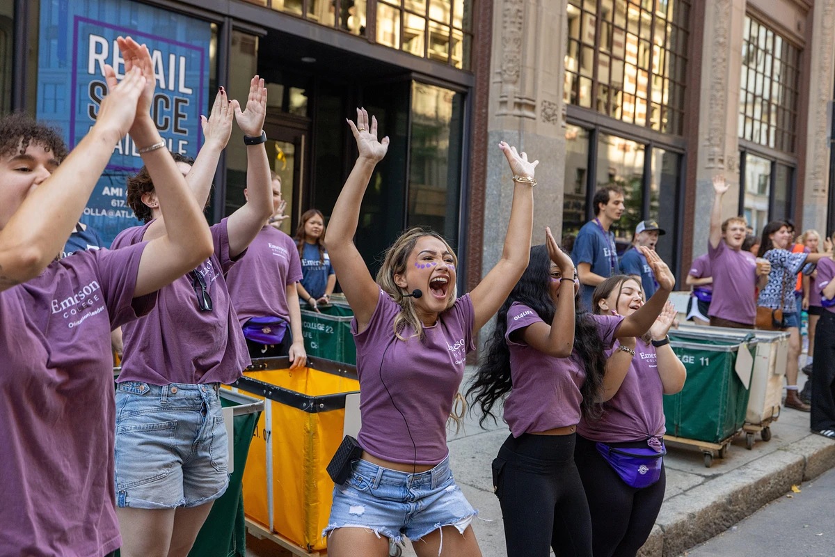 A group of enthusiastic people in matching purple shirts are cheering and raising their arms outdoors.