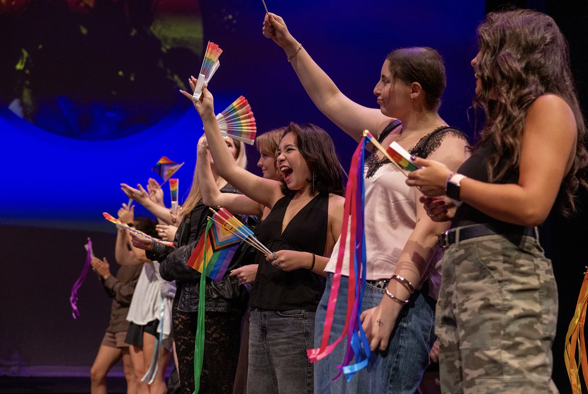 A group of people on stage enthusiastically wave rainbow flags and ribbons.