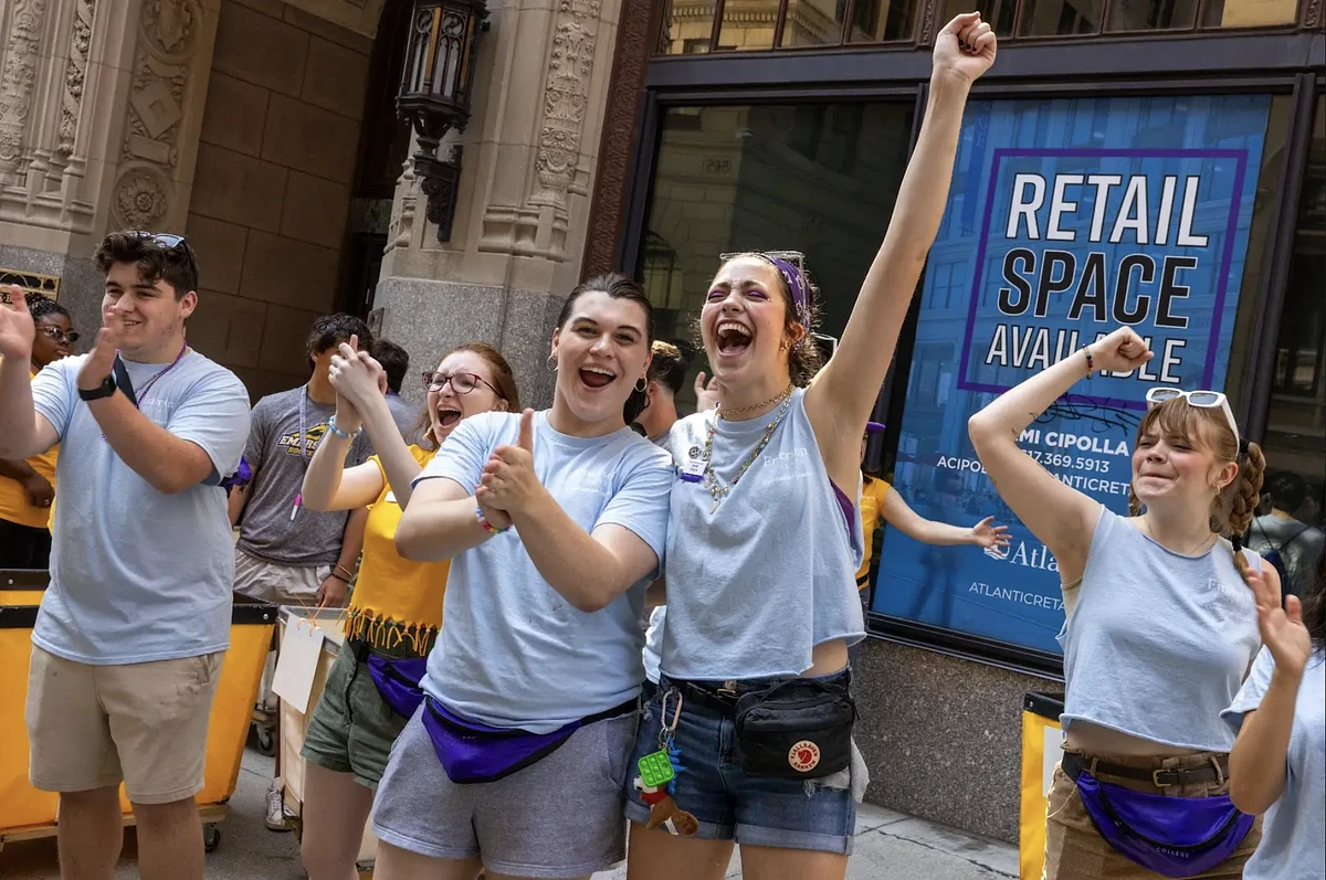 A group of people enthusiastically cheers and claps in front of a building with a "Retail Space Available" sign.